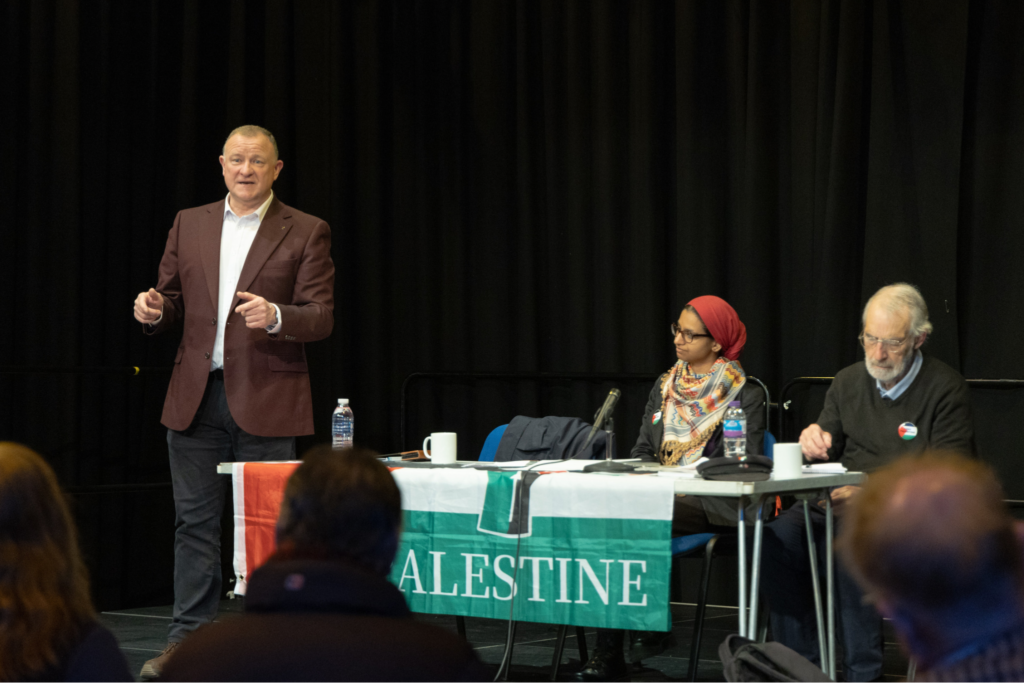 Drew Hendry MP stood up, speaking to audience at Solidarity Gathering. He is stood up next to the other two panel members who are sat down. The table that forms the panel has a Palestinian flag draped over it and reads "PALESTINE" in large white letters. The backdrop is a black curtain. 