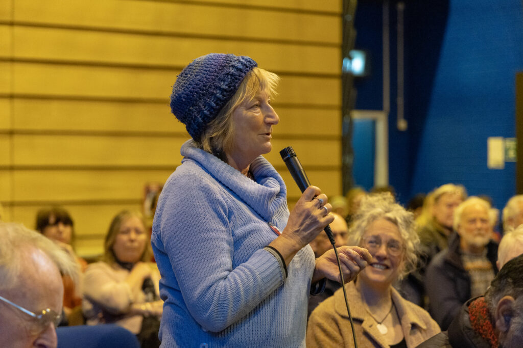 An audience member is standing looking towards the panel, with the rest of the audience sat around them. The audience member is wearing a light blue jumper and a blue hat. Another member of the audience is looking up at them and smiling. 