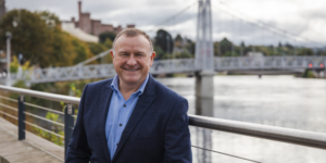 image of Drew Hendry MP by the River Ness in Inverness. Inverness Castle can be seen over his shoulder